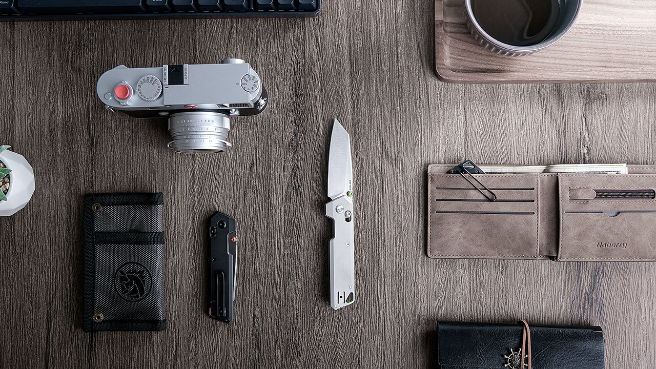 Elegant desk setup featuring two folding knives alongside professional accessories including a Leica camera, leather wallet, coffee cup, and keyboard on a wooden desk surface, representing a sophisticated office environment