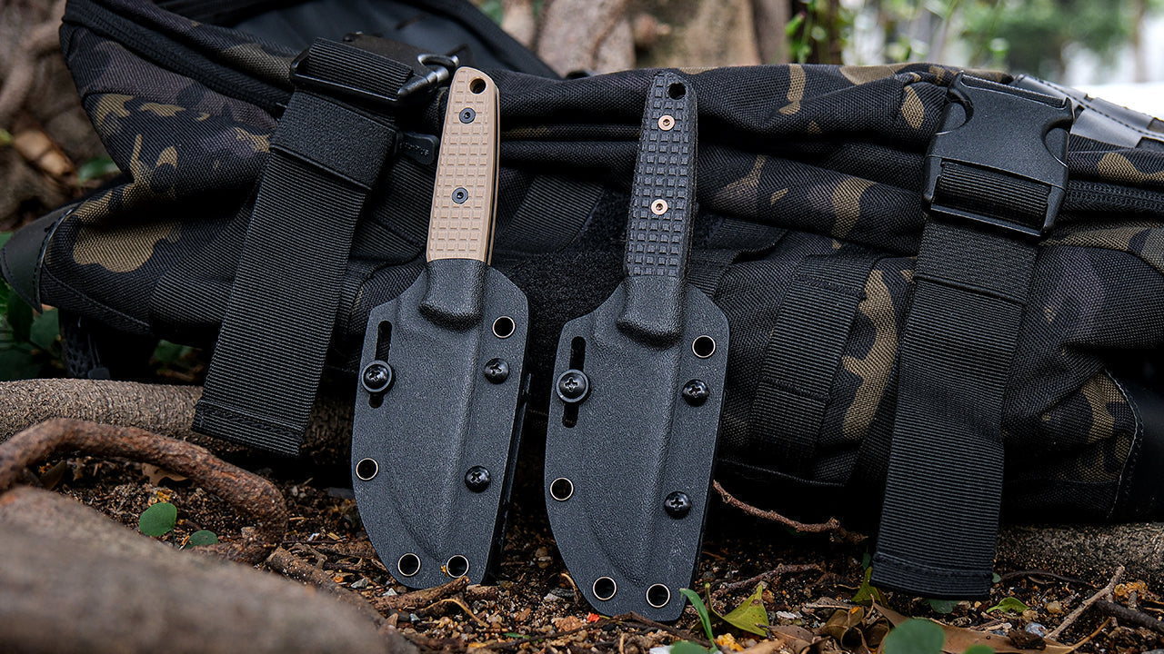 Two fixed-blade knives, one with a tan handle and one with a black handle, are shown in their black Kydex sheaths, leaning against a camouflage backpack in an outdoor setting