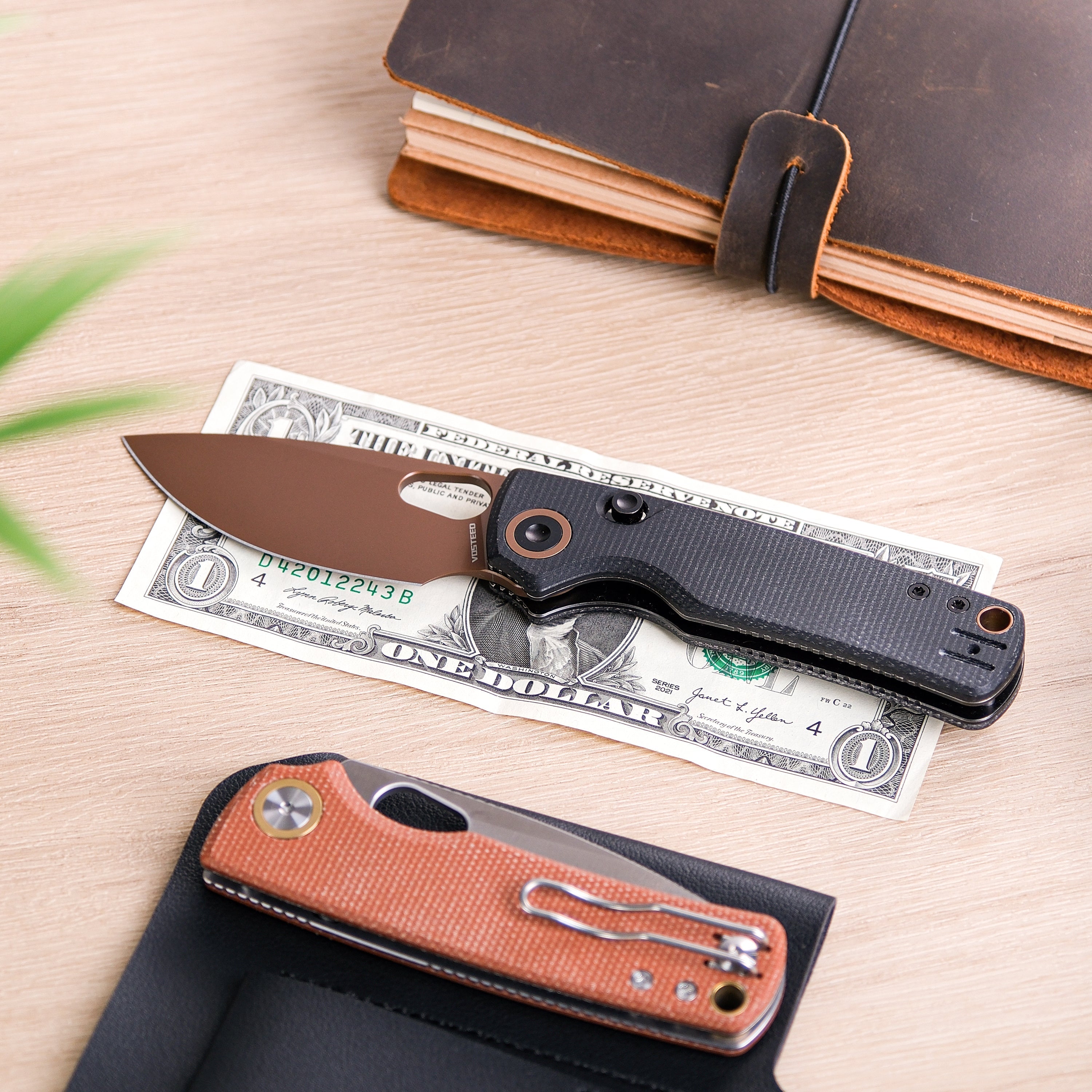 Folding knife with a brown blade and black handle placed on a one-dollar bill next to a notebook and another folding knife in a leather case
