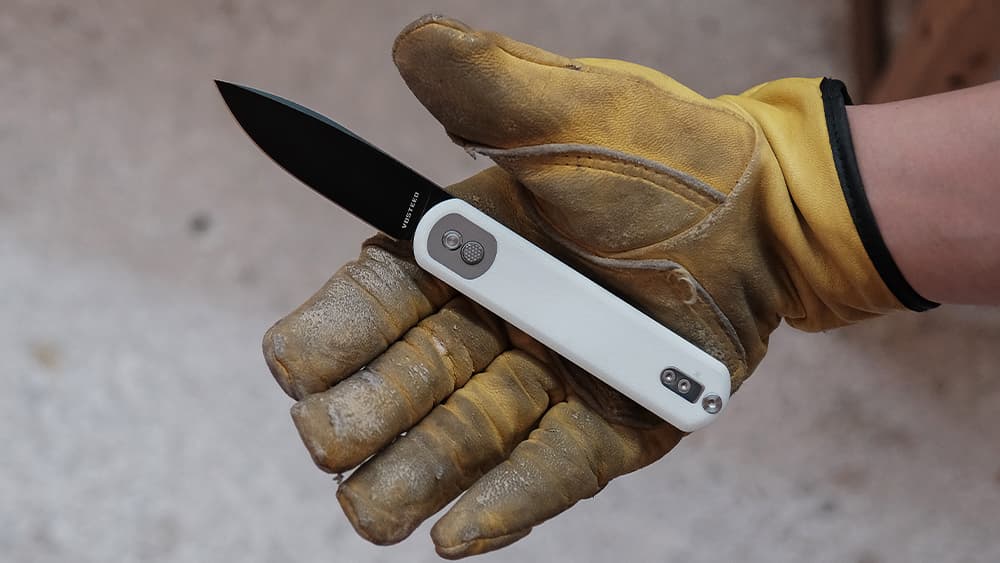 A gloved hand holding an open folding knife with a black blade and white handle, showcasing the knife's compact design and sharp edge against a light gray background