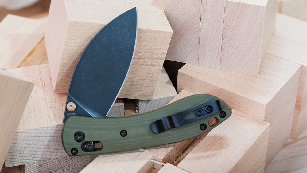 An open folding knife with a dark blade and olive green handle resting on a pile of wooden blocks or lumber pieces, displaying its tactical design and cutting edge