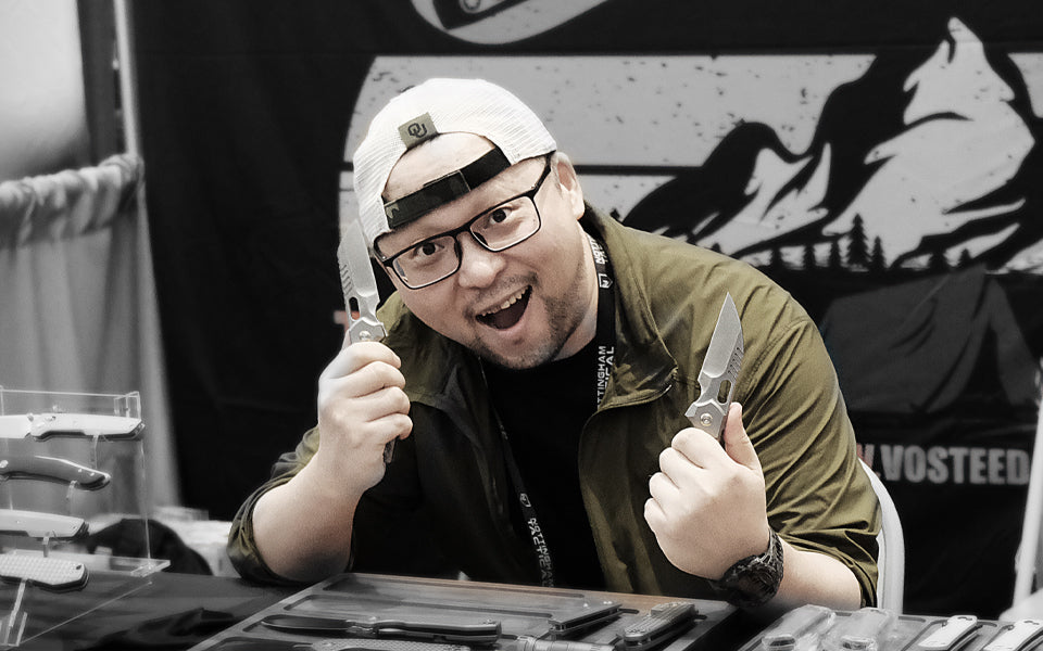 Designer Yue Dong at the Vosteed knife display booth, wearing a white baseball cap and glasses, enthusiastically showcasing two refined folding knives. The background features a Vosteed brand wall decorated with mountain silhouette patterns