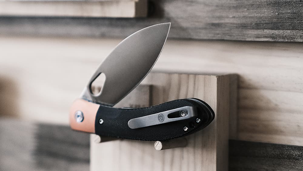 Folding knife with a drop point blade in satin finish, featuring a black G10 handle with copper backspacer, displayed partially open on a wooden surface