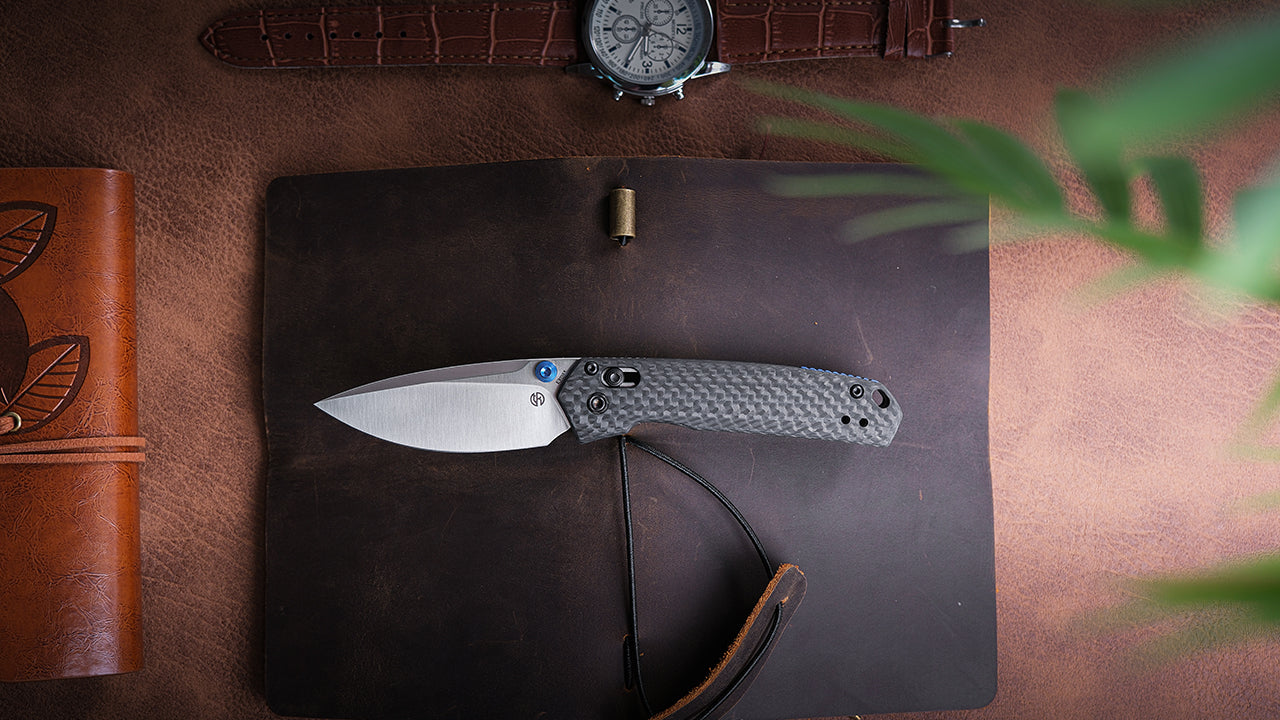 An overhead shot of a folding knife with a silver blade and carbon fiber handle resting on a brown leather surface, next to a notebook and a watch