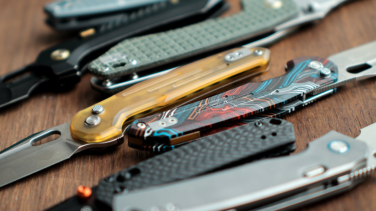 Multiple folding knives arranged side by side on a wooden surface, showcasing various handle materials including black, gray, and white, demonstrating the appearance of modern EDC knives
