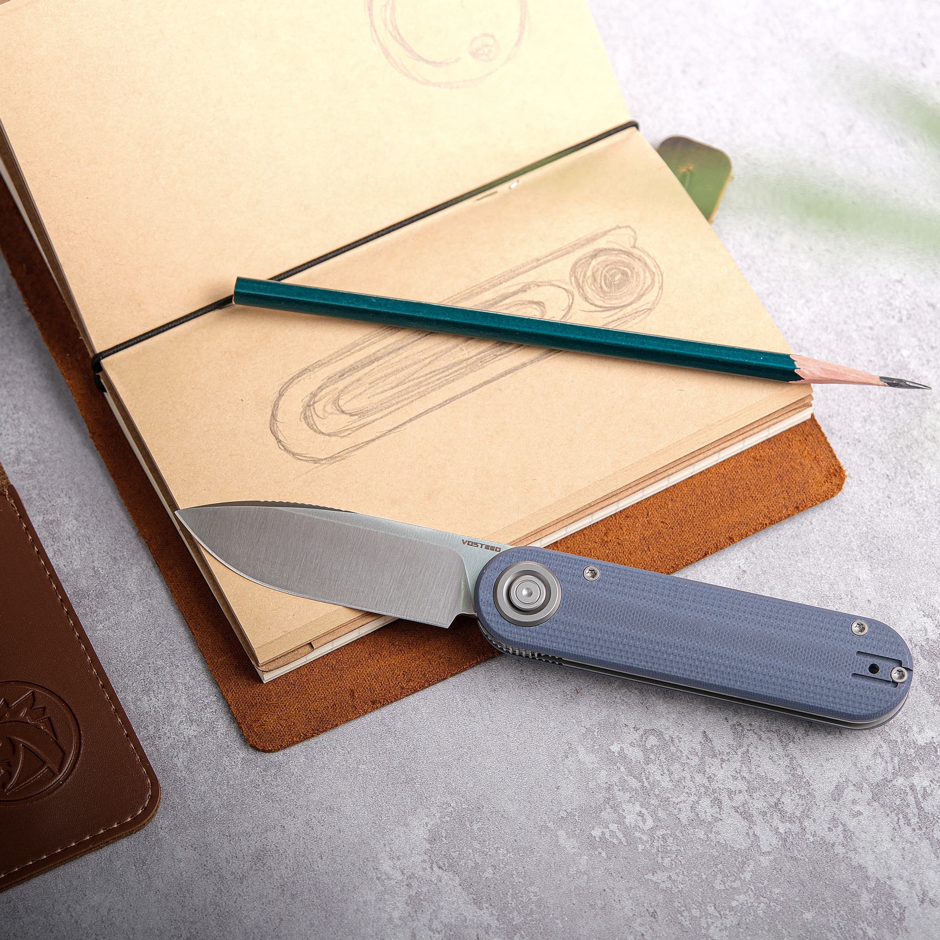 Folding knife with a blue textured handle and stainless steel blade resting on a sketchbook with pencil drawings of knife designs. A green pencil is placed across the open notebook
