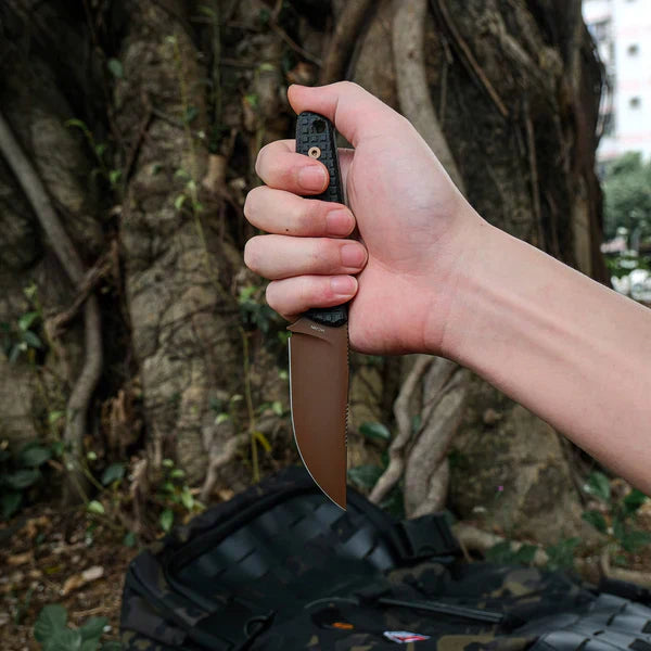 Hand holding a compact bushcraft knife with a black textured handle and brown blade against a forest backdrop with tree roots visible